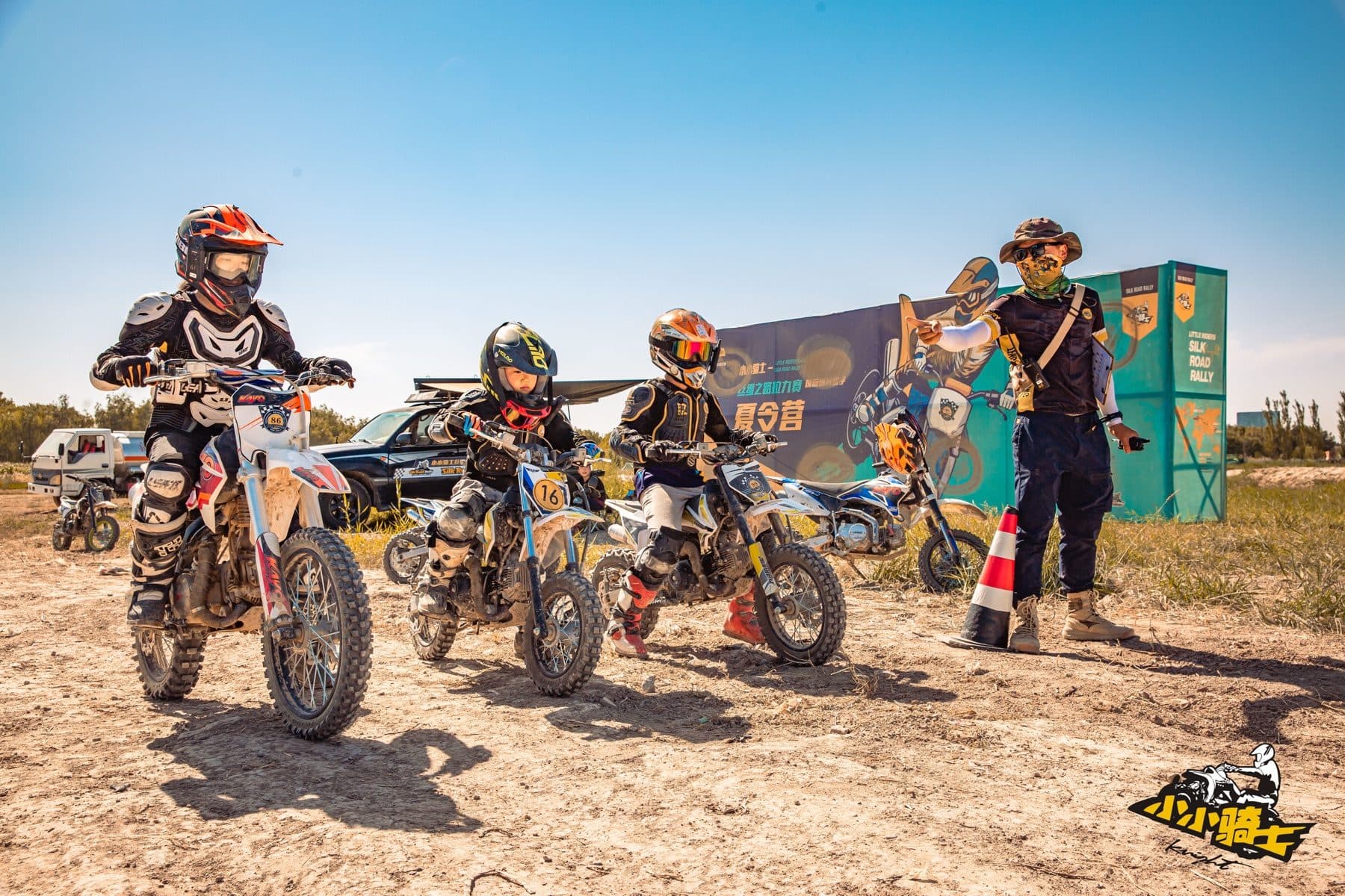 Children in protective equipment waiting on off-road motorcycles while an adult guide gives instructions